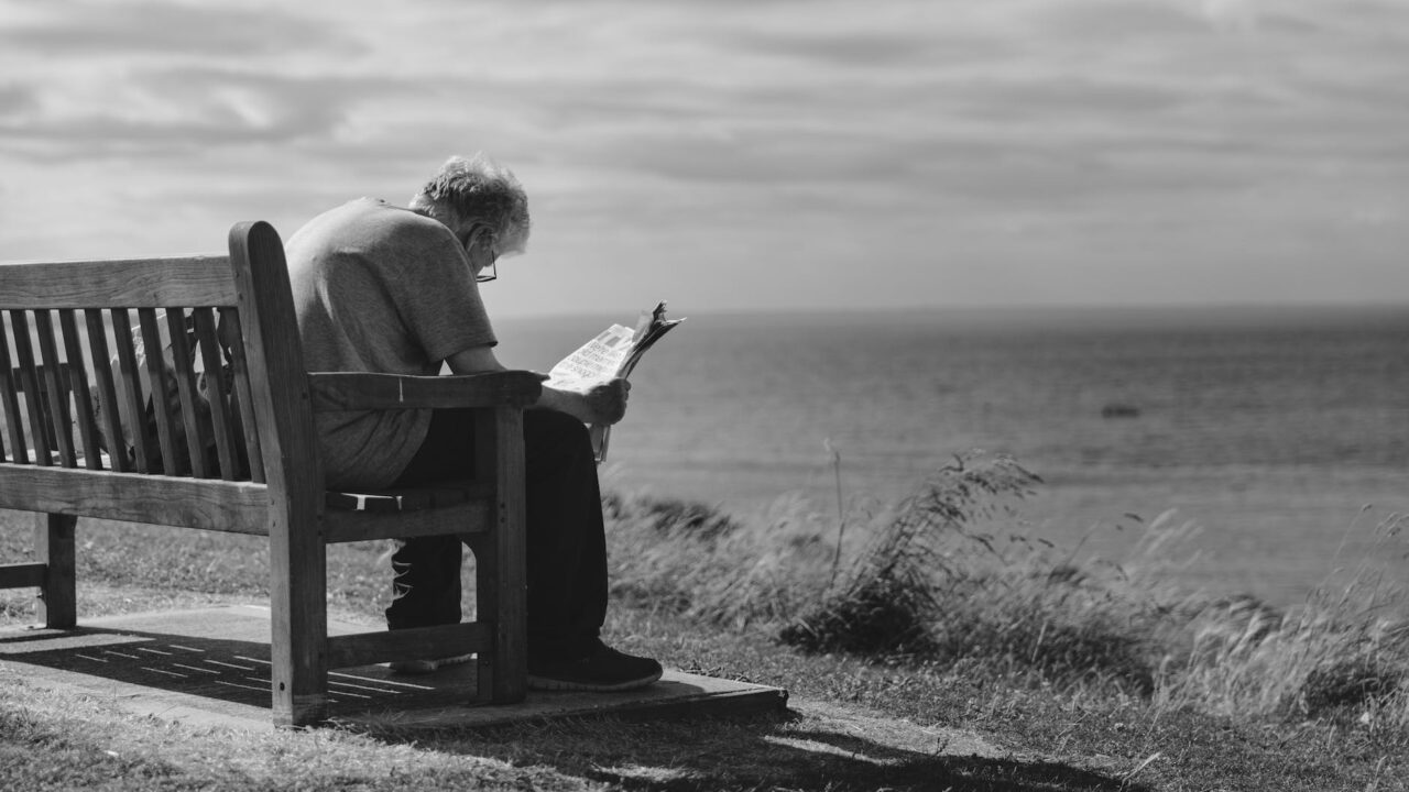 grayscale photo of man sitting on brown wooden bench reading news paper during day time